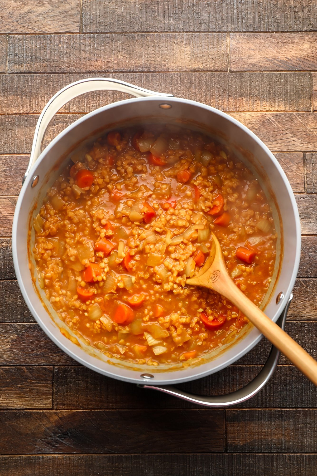 using a wooden spoon to stir carrot and lentil soup in a large grey pot.