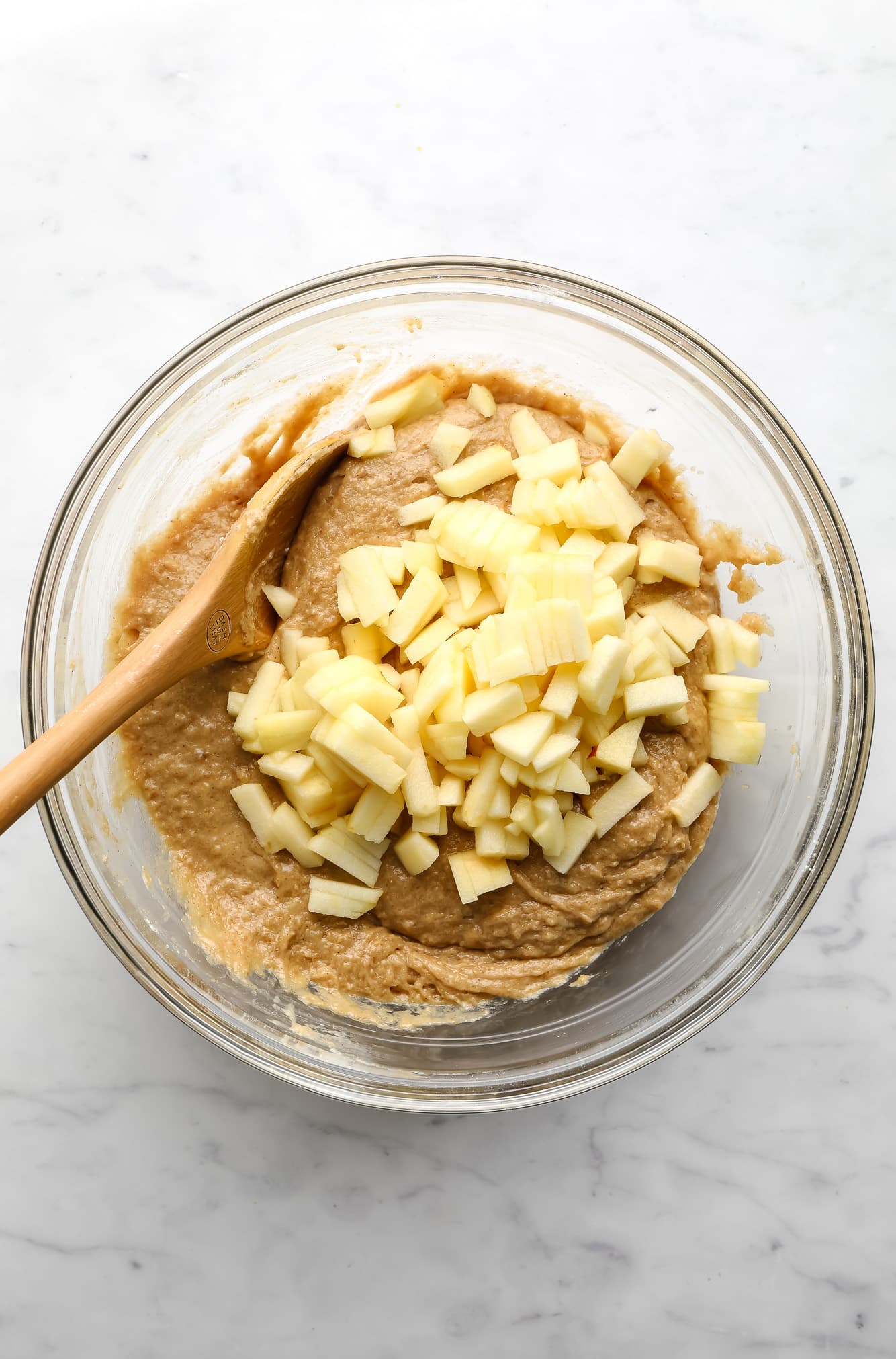 using a wooden spoon to stir chopped apples into vegan apple muffin batter in a glass bowl.