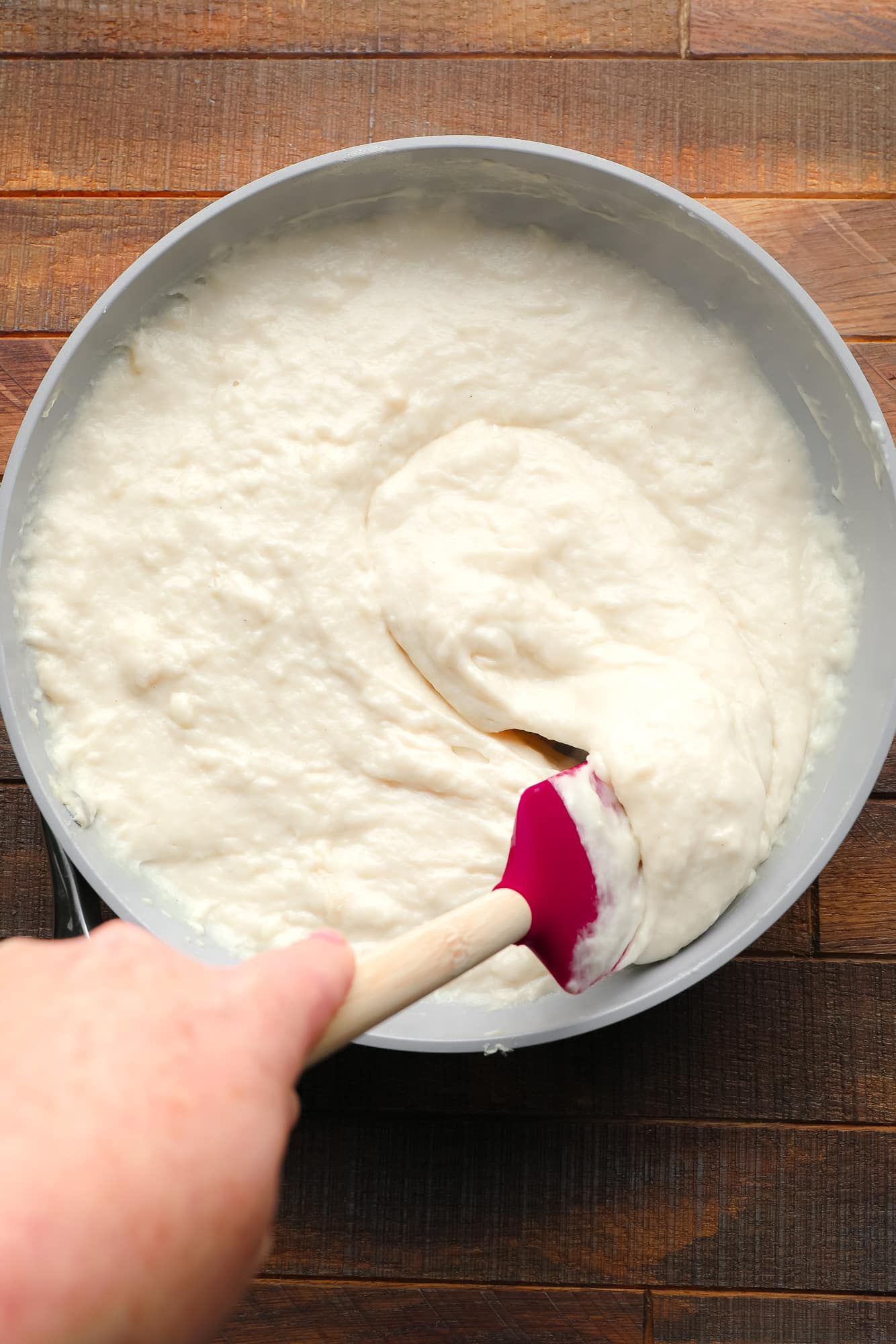 using a red spatula to stir creamy vegan brie cheese in a grey saucepan.