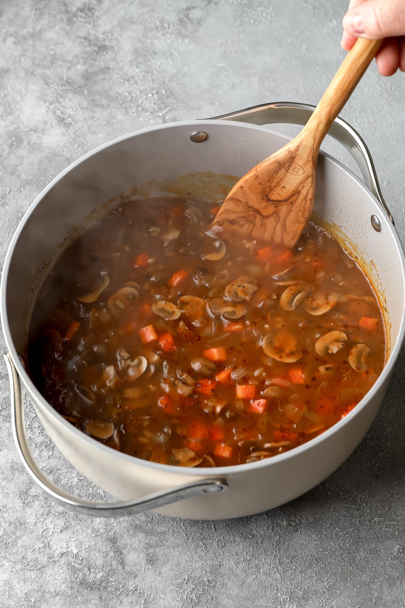 using a wooden spoon to stir mushroom soup in a large grey pot.