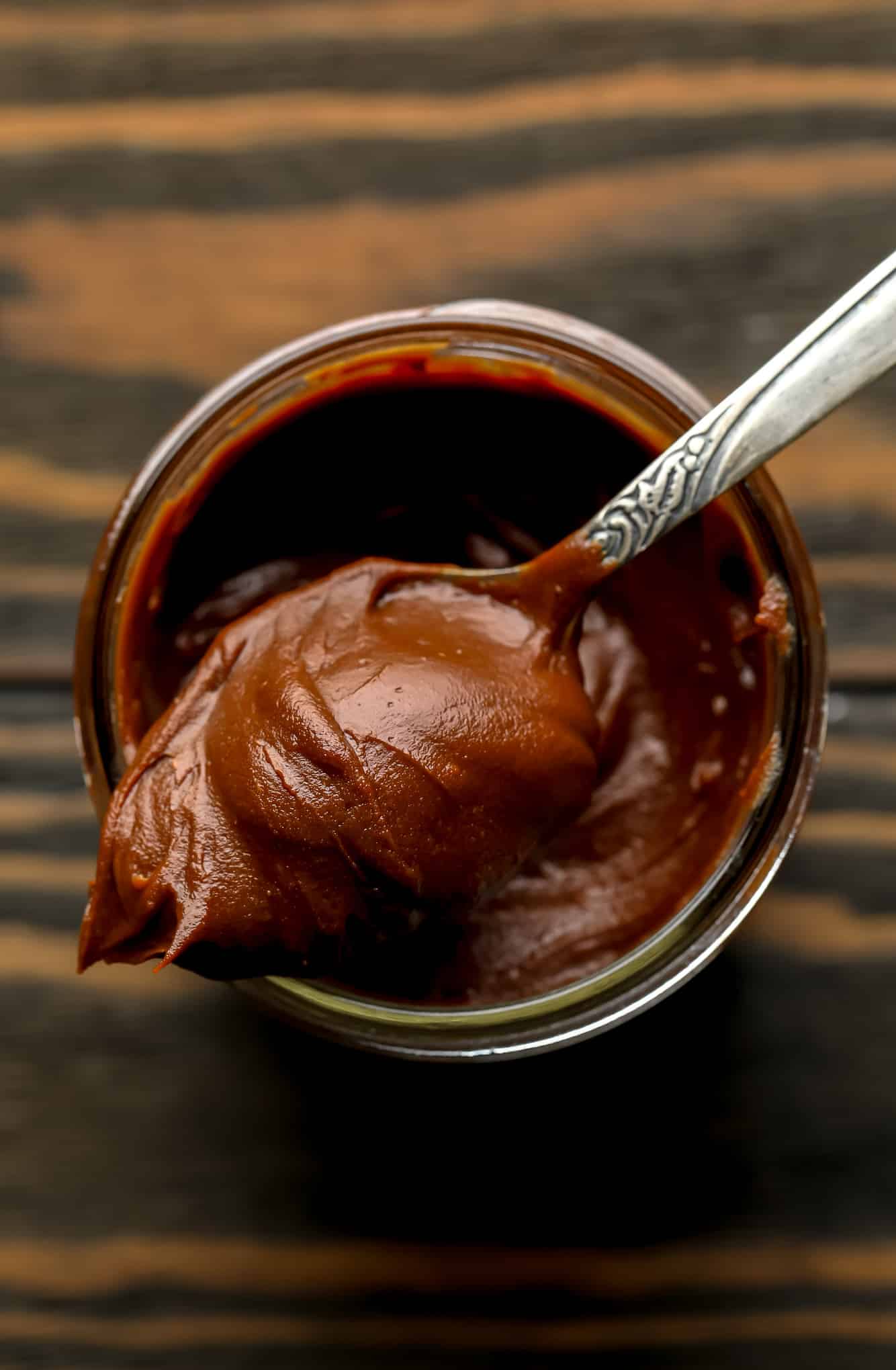 a spoon with a scoop of vegan nutella on top of a glass jar filled with more nutella.