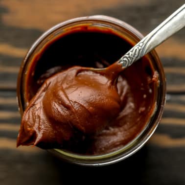 a spoon with a scoop of vegan nutella on top of a glass jar filled with more nutella.