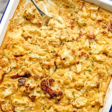 using a large metal spoon to scoop a spoonful of vegan cauliflower cheese from a large baking dish.