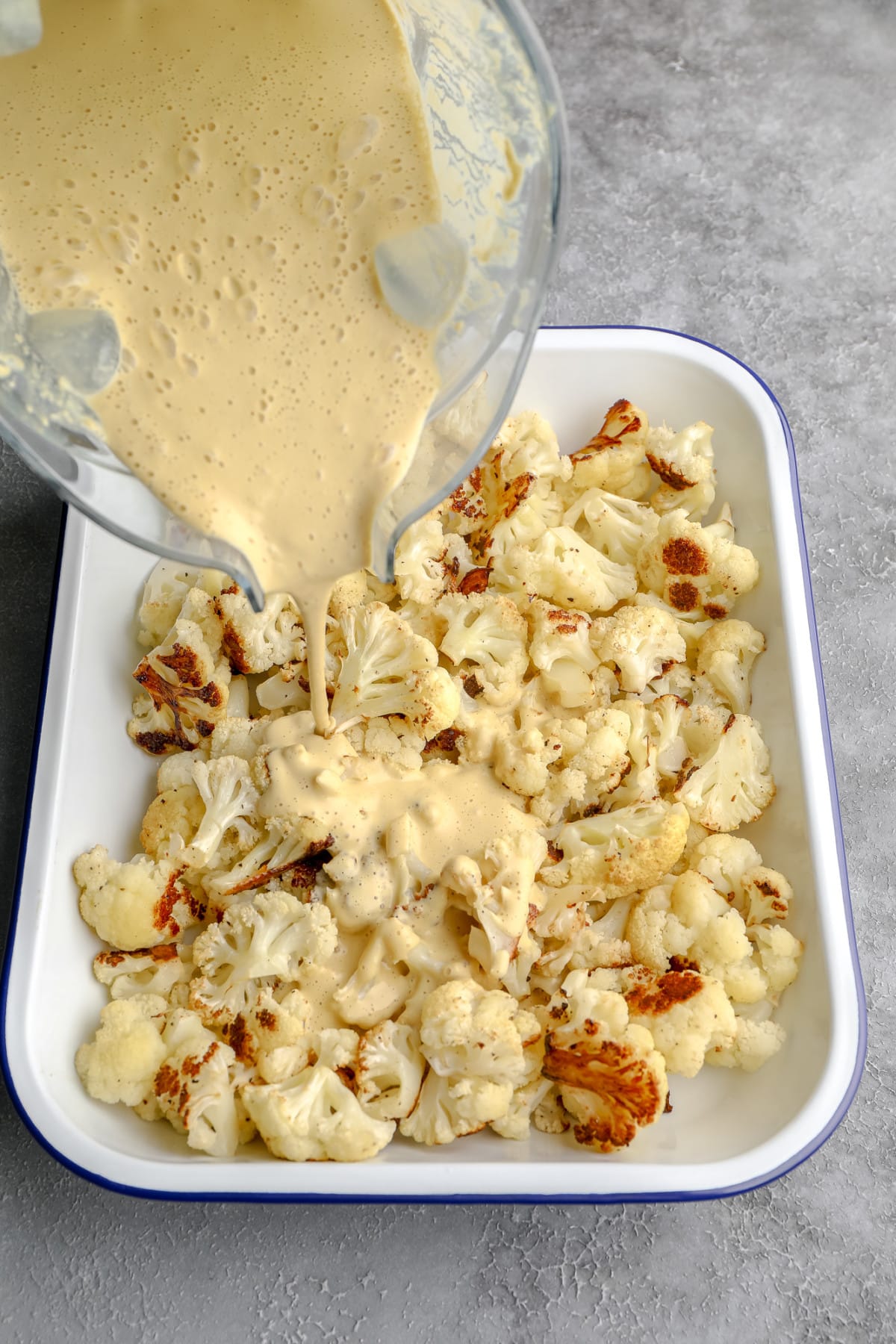 pouring vegan cheese sauce over roasted cauliflower florets in a large white baking dish.