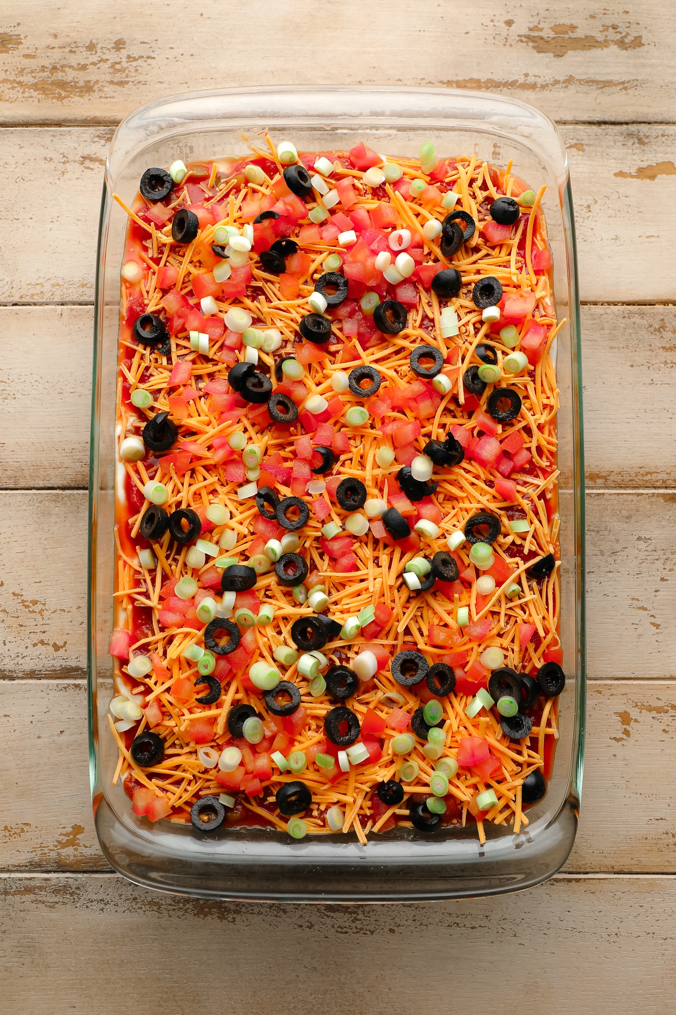 Vegan 7 Layer Dip topped with black olives, shredded vegan cheese, and diced tomatoes in a glass casserole dish.