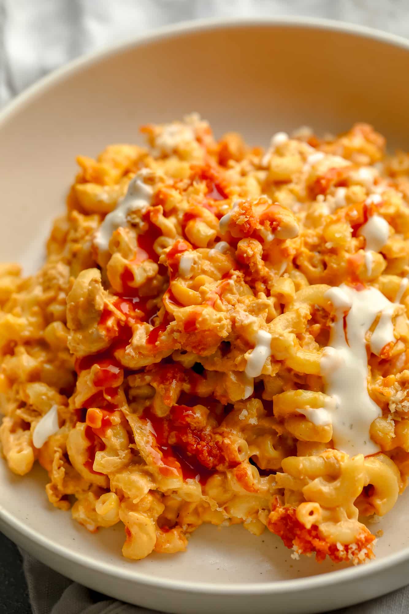 close up on Vegan Buffalo Mac and Cheese topped with white vegan ranch and hot sauce in a beige bowl.