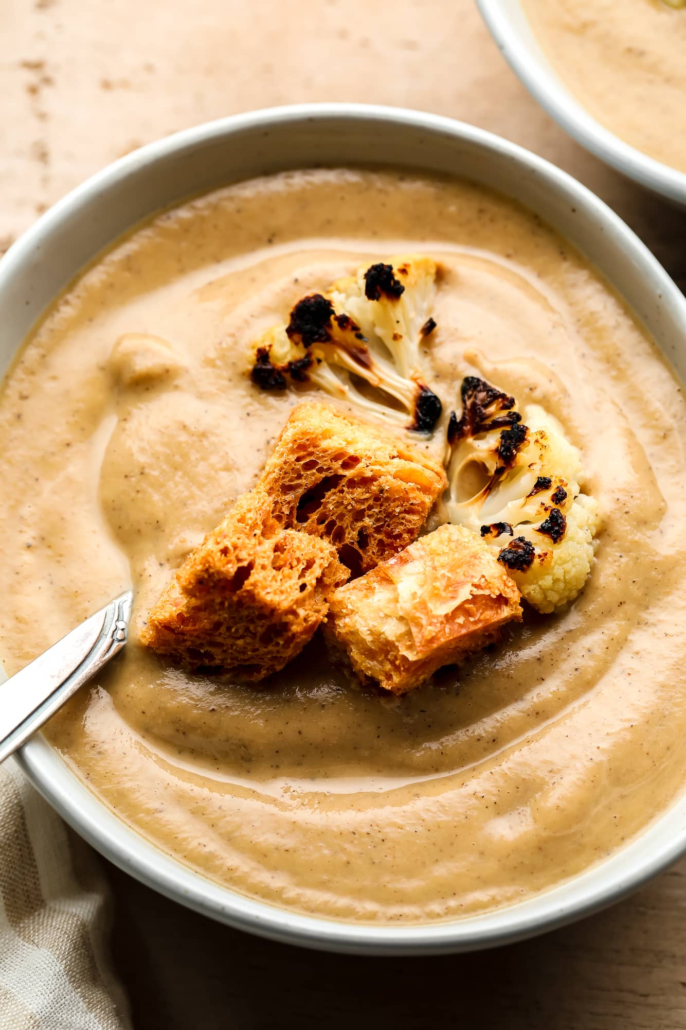 close up on roasted cauliflower soup topped with croutons and charred cauliflower florets in a white bowl.