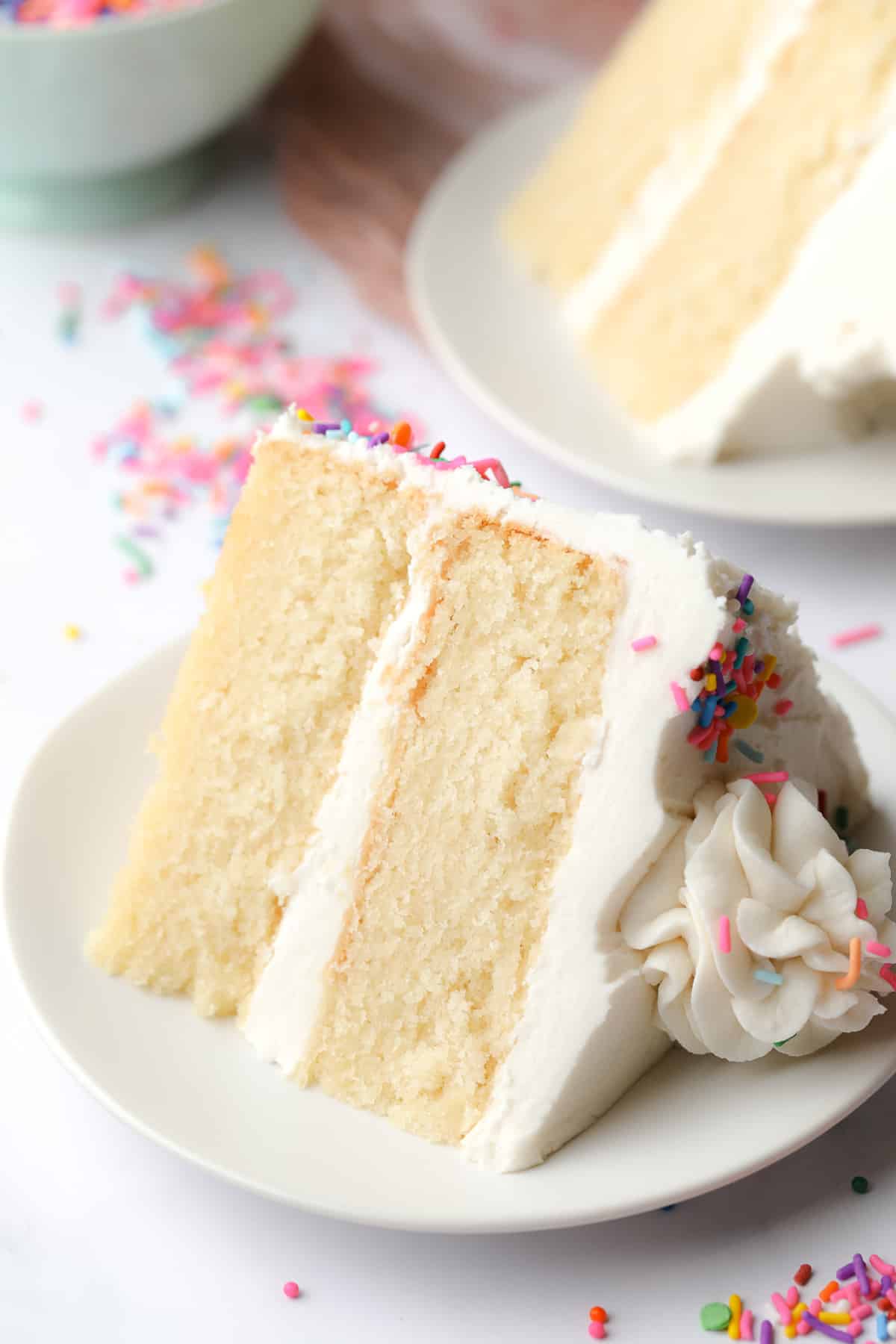 vegan vanilla cake on a white plate, another piece in the background, showing fluffy texture and white frosting with sprinkles outside