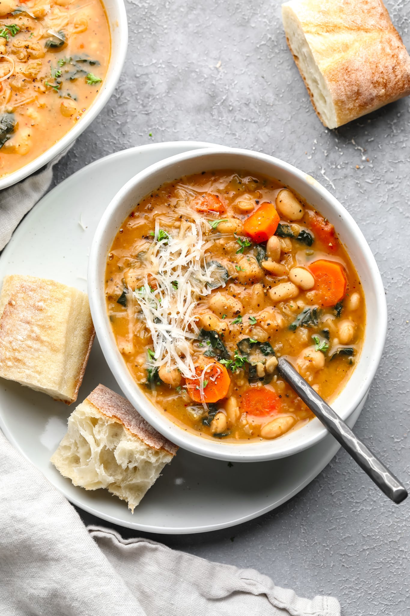 a white bowl full of white bean soup topped with shredded vegan parmesan and pieces of bread on the side.