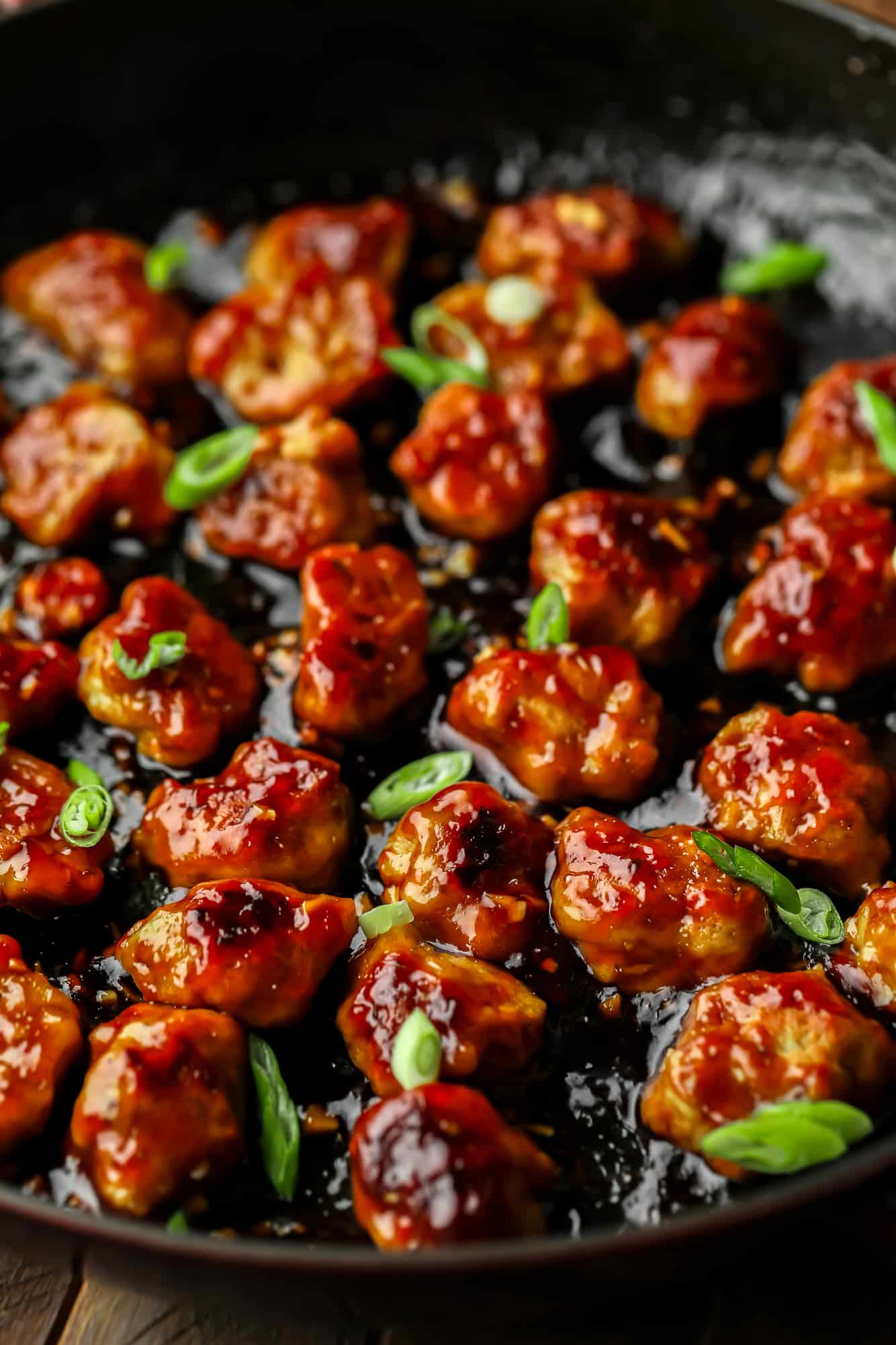 close up on bite-sized seitan nuggets covered in a "honey" ginger sauce and cooking in a large black skillet.