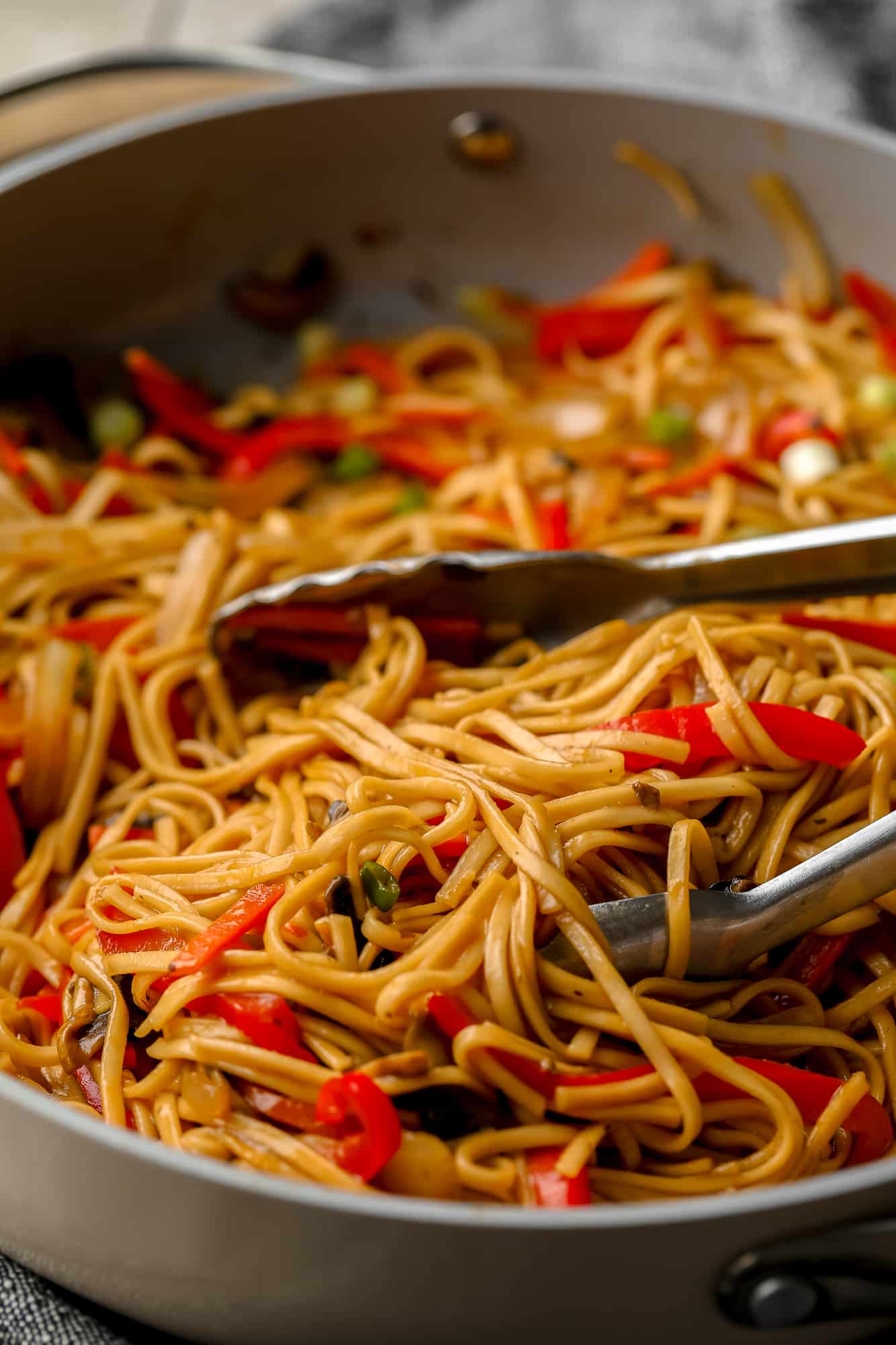 metal tongs in a batch of vegan lo mein in a large gray skillet.