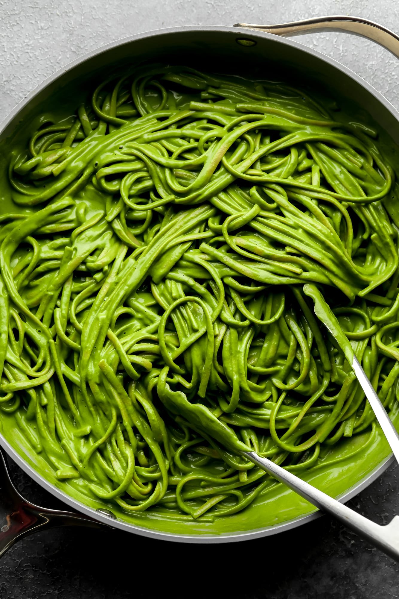 using metal tongs to toss long pasta noodles in a bright green sauce in a large grey pot.