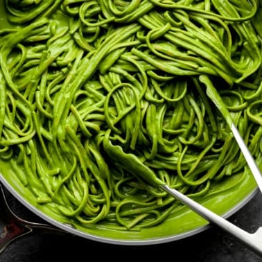 using metal tongs to toss long pasta noodles in a bright green sauce in a large grey pot.