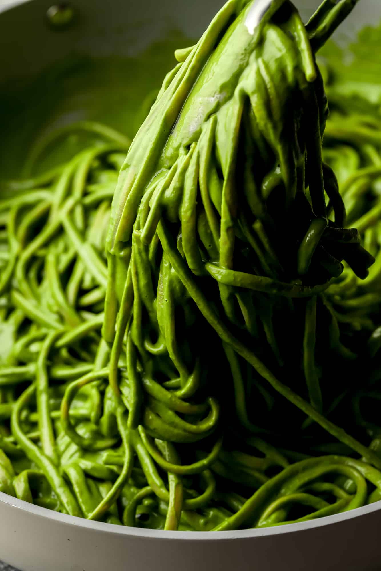 metal tongs lifting a scoop of green pasta from a large pot.