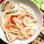 hummus topped with paprika and oil in a white bowl with pita bread on the side.