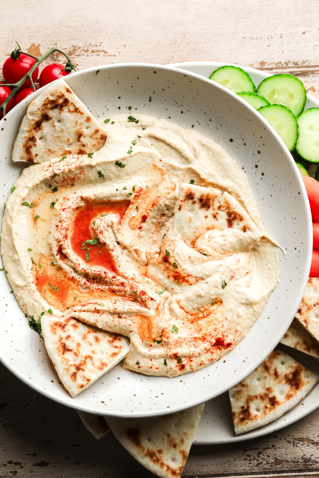 hummus topped with paprika and oil in a white bowl with pita bread and vegetables on the side.