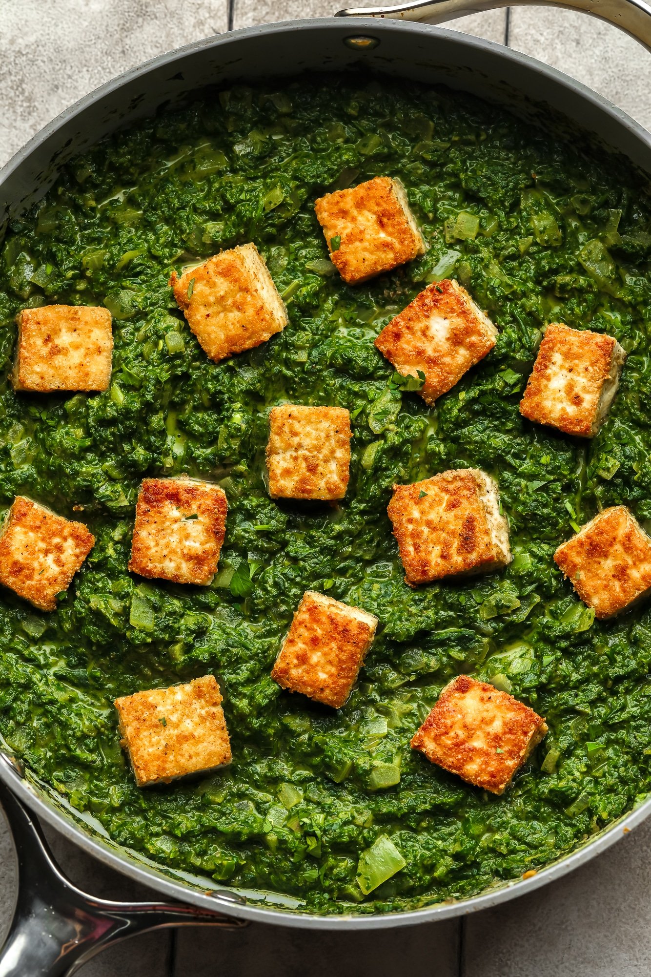 golden tofu cubes on top of a batch of vegan palak paneer in a large skillet.
