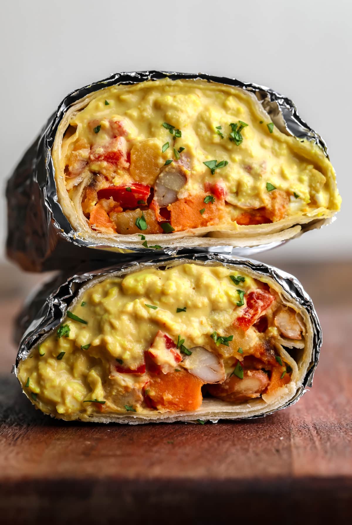 burrito cut in half with foil around it on wood board with eggy mixture plus veggies