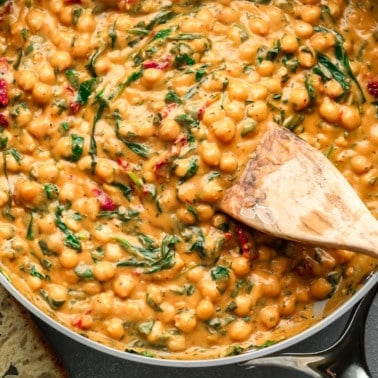 close up on a wooden spoon stirring a batch of Marry Me Chickpeas in a large skillet.