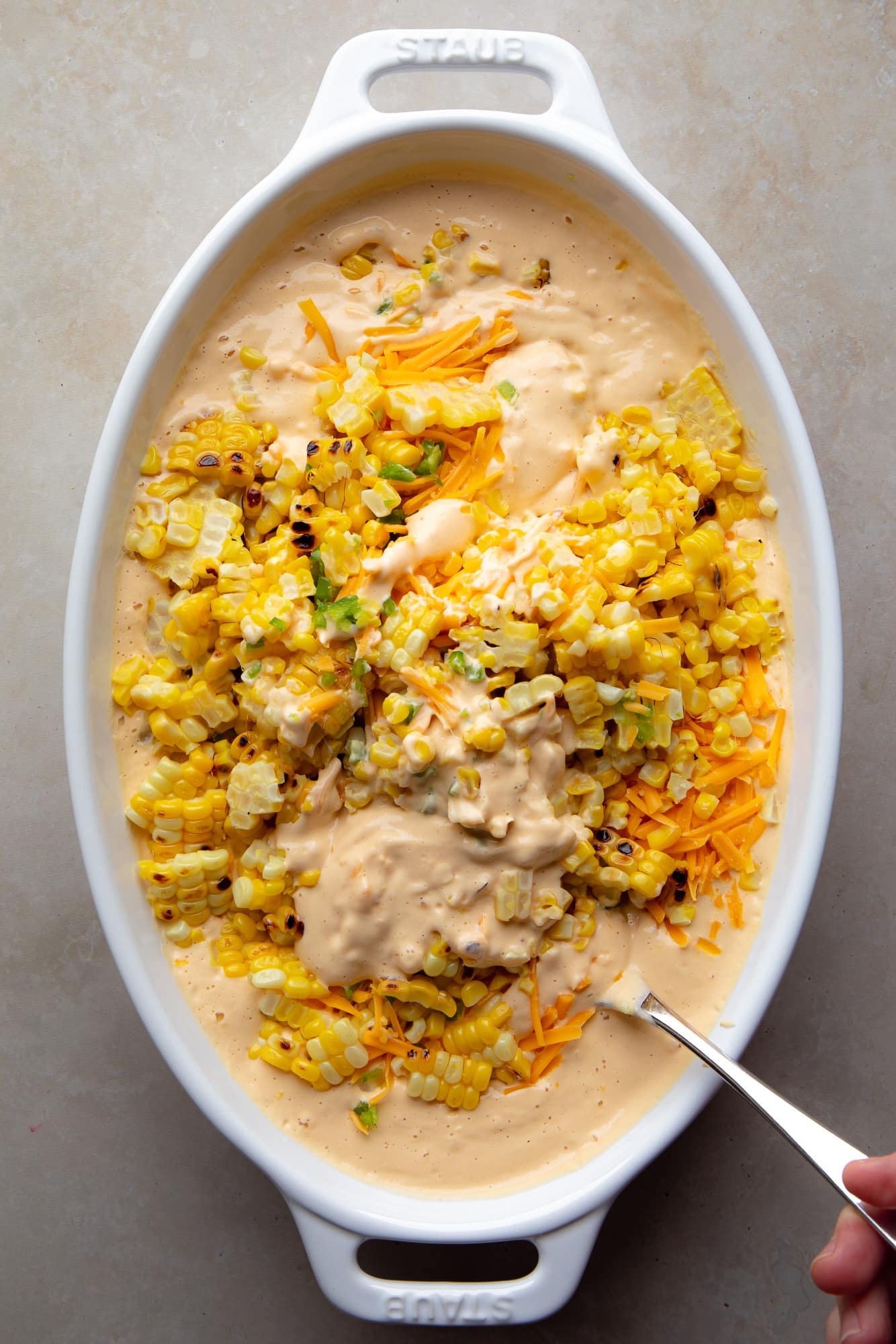 using a spoon to combine the ingredients for vegan Mexican street corn dip with a creamy cashew sauce in a baking dish.