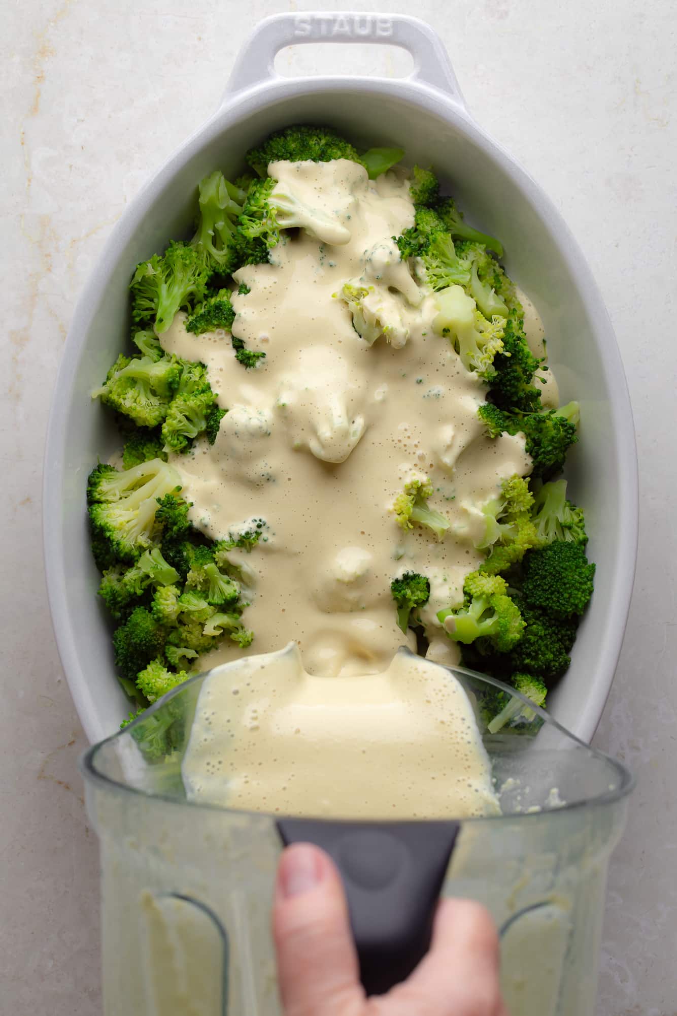 womans hand pouring vegan cheese sauce over broccoli in a large casserole dish.