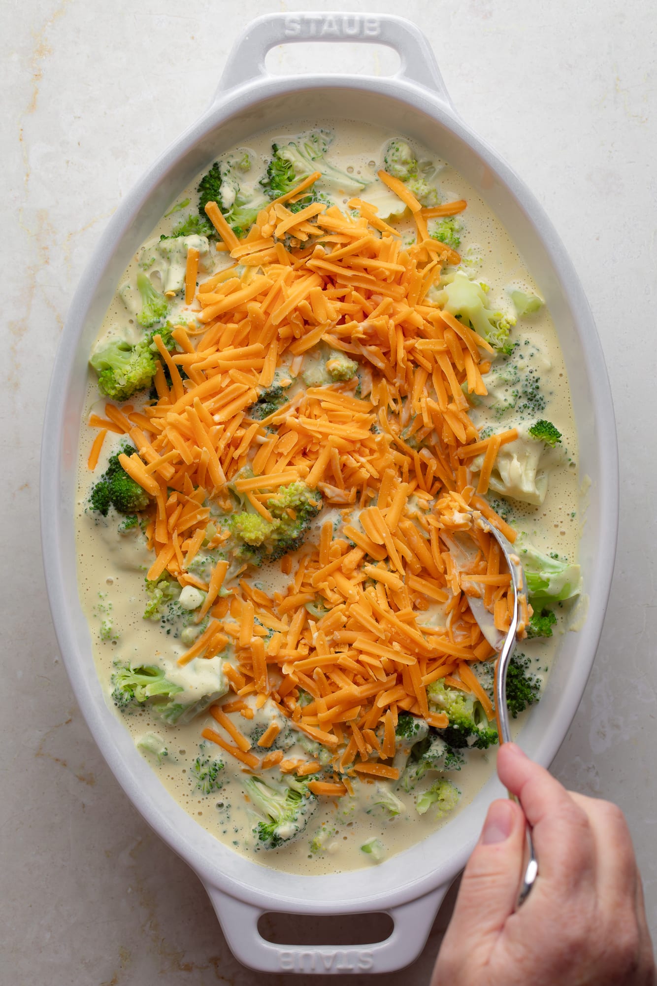 womans hand using a spoon to stir vegan cheese shreds into a vegan broccoli casserole.