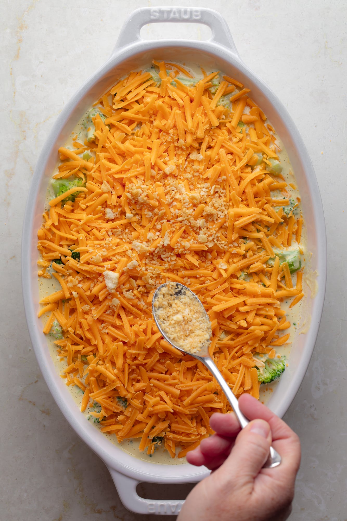 womans hand sprinkling breadcrumbs over a vegan cheese-topped vegan broccoli casserole.