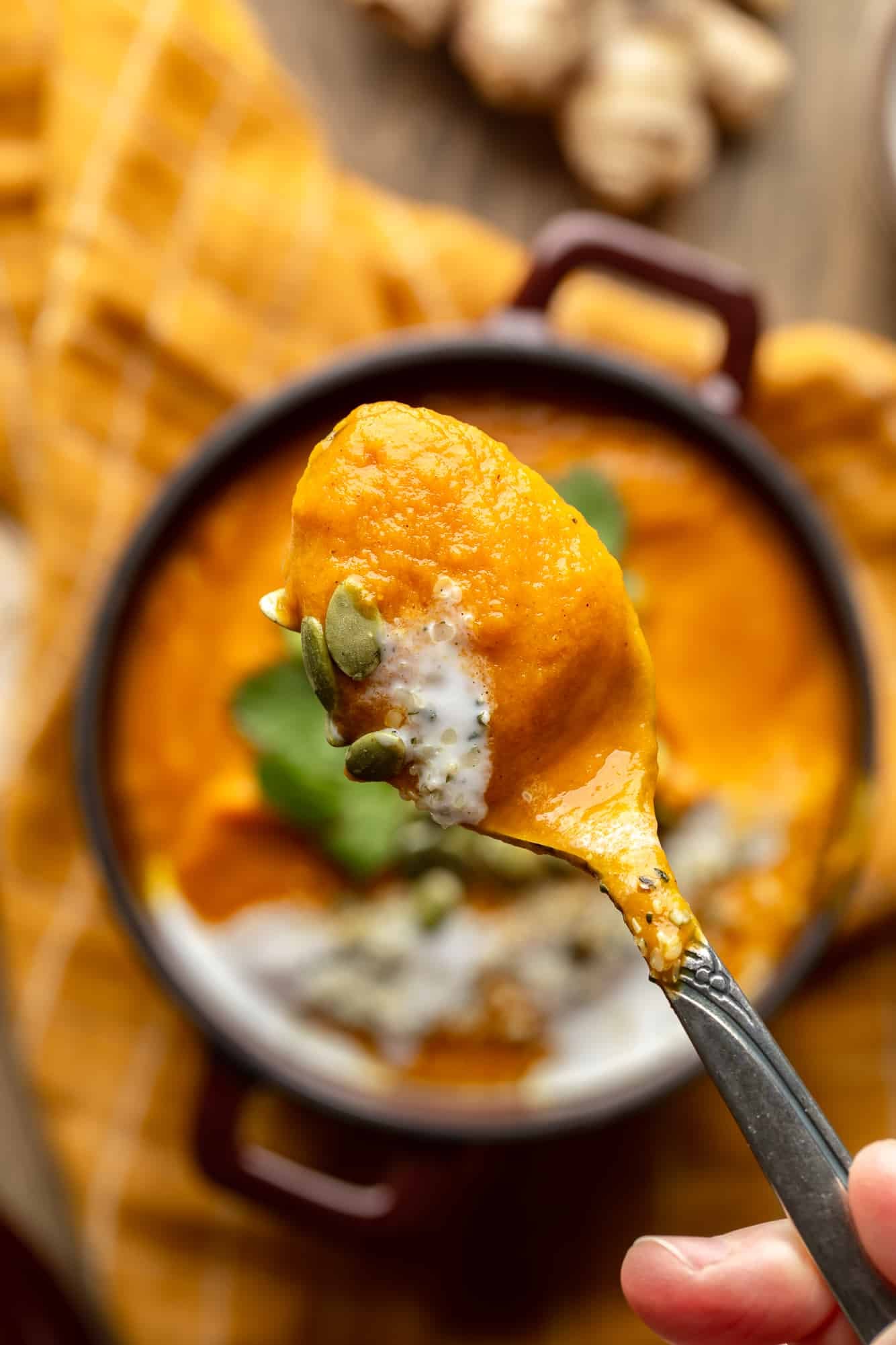 close up on a spoonful of vegan carrot and ginger soup with a bowl of the soup in the background.