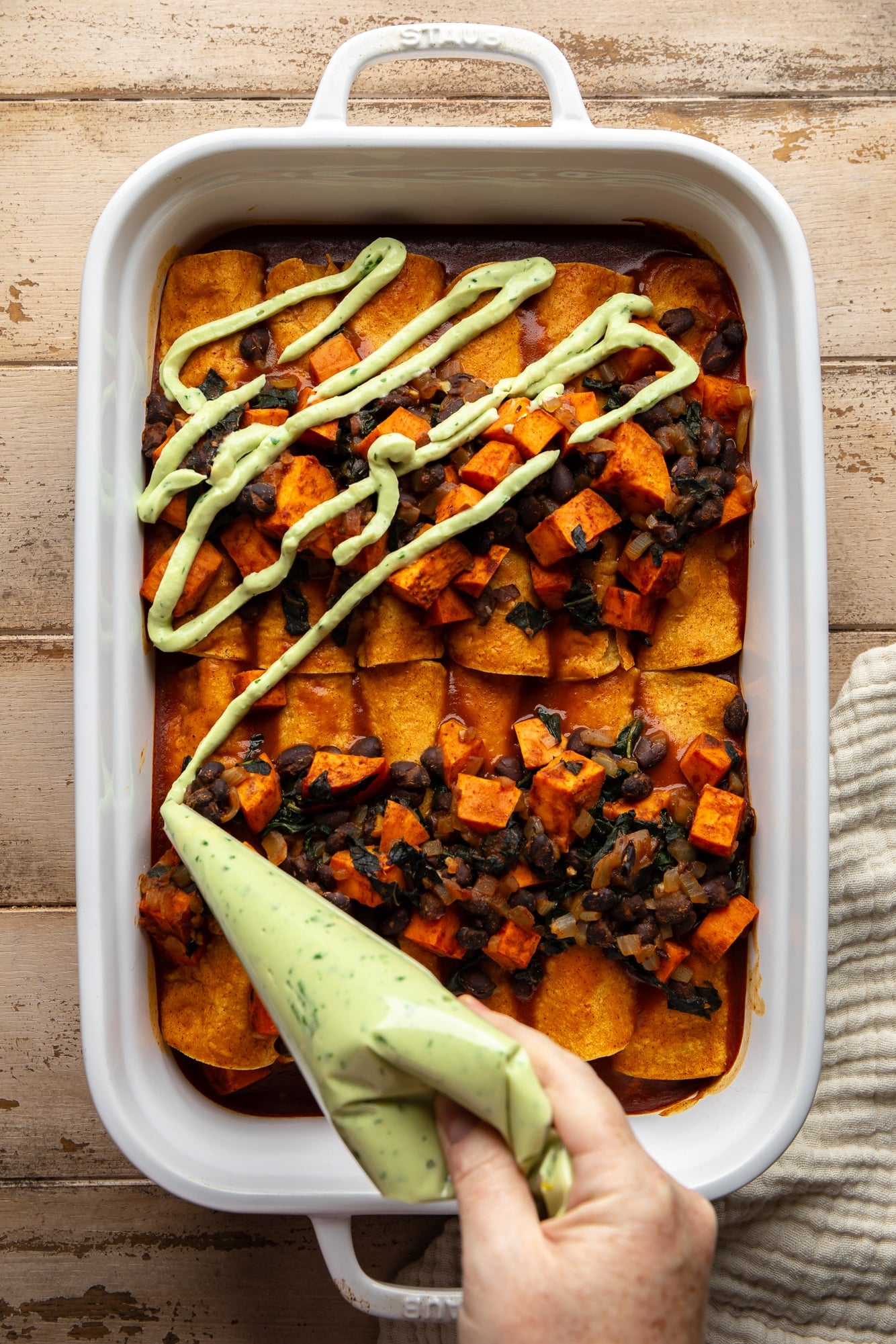 a womans hand piping vegan avocado crema on top of sweet potato black bean enchiladas in a large casserole dish.
