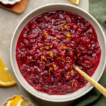 Overhead view of a bowl of orange cranberry sauce topped with orange zest with orange slices and fresh cranberries on the side.