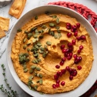 overhead view of a bowl of orange pumpkin hummus topped with pumpkin seeds and pomegranate seeds.