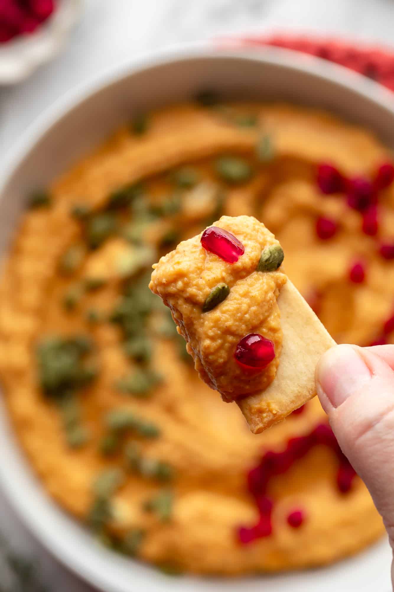 close up on a womans hand holding a pita chip covered in pumpkin hummus.