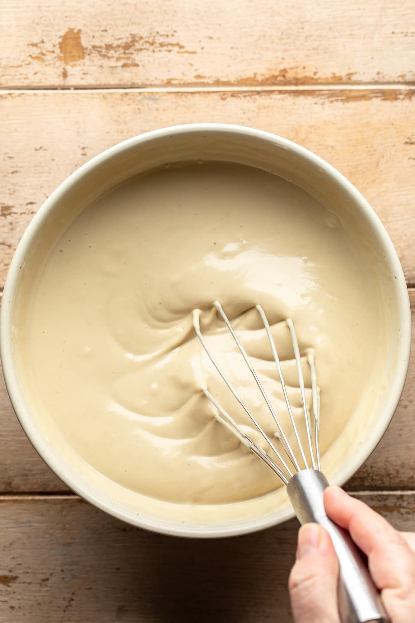 a womans hand whisking garlic tahini sauce in a large white bowl.