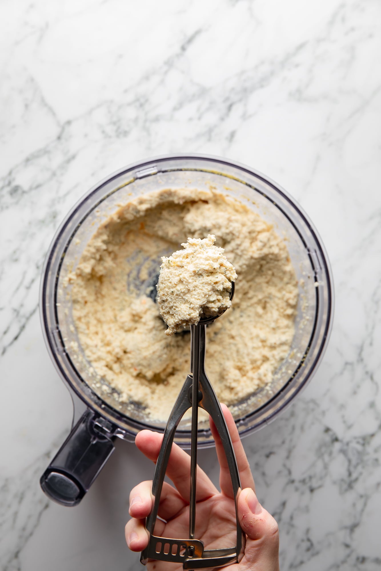 a womans hand holding a cookie scoop filled with a tofu meatball mixture above a food processor.