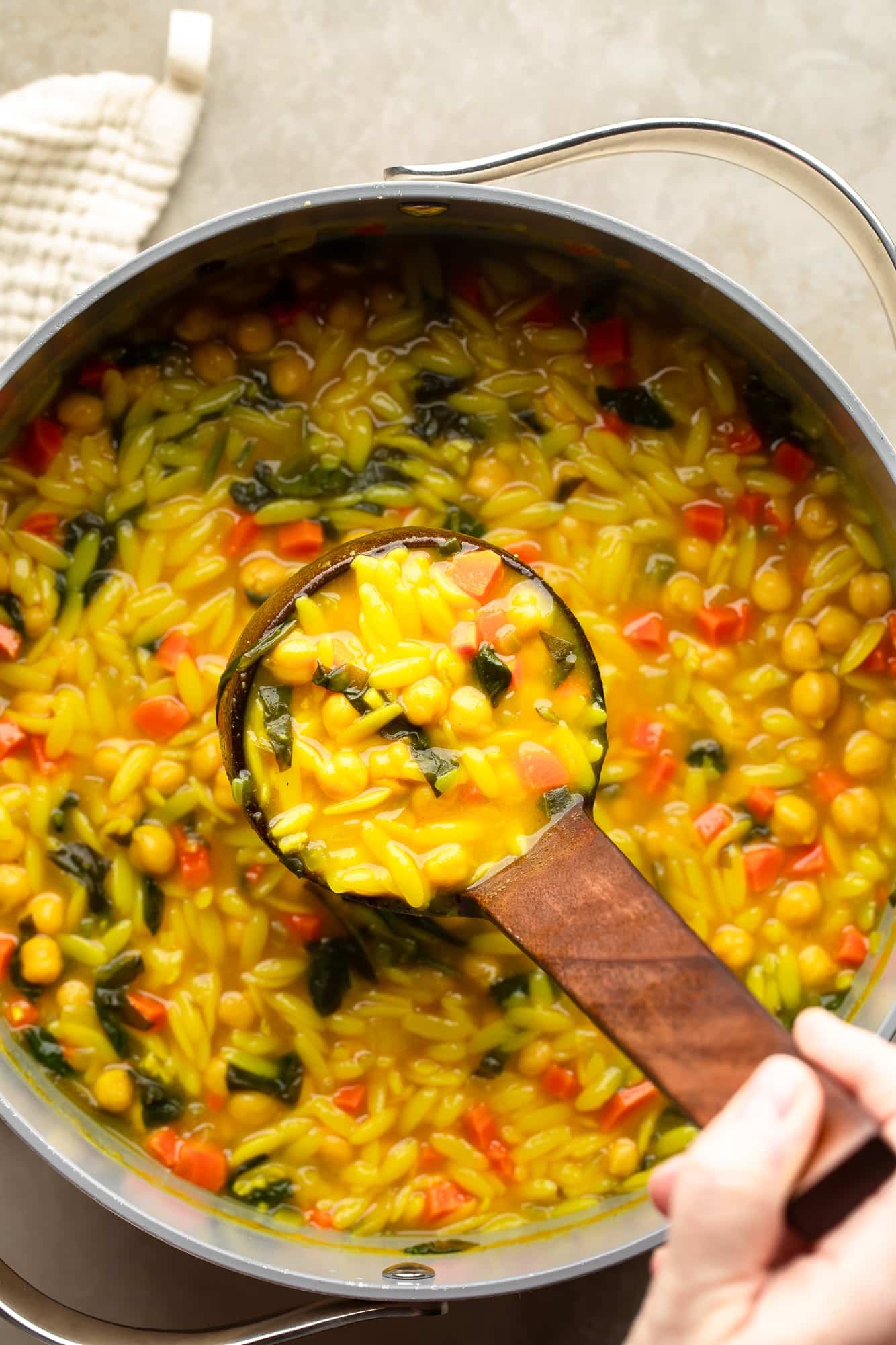 overhead view of a hand lifting a ladle out of a large pot of Golden Orzo Soup with Chickpeas.