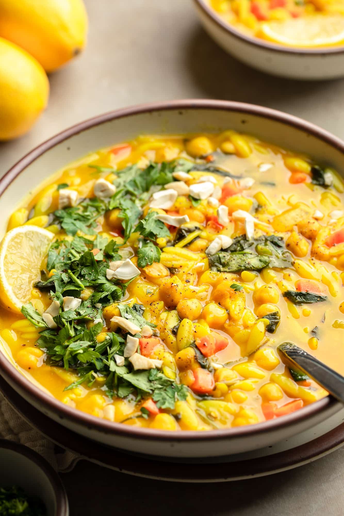 side view of a bowl of Golden Orzo Soup with Chickpeas topped with a lemon wedge, green herbs, and nuts.