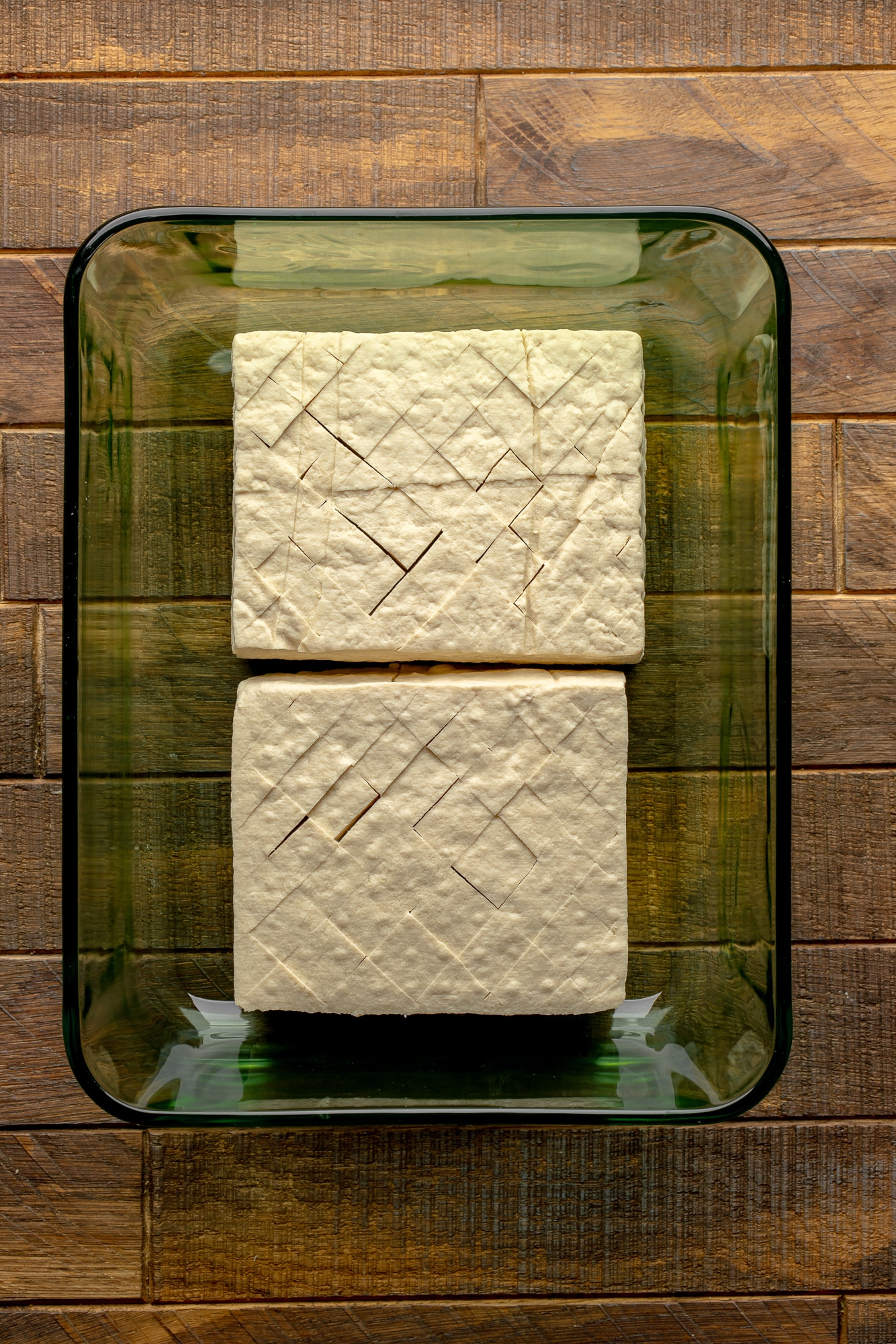 overhead view of 2 scored tofu blocks in a glass container.