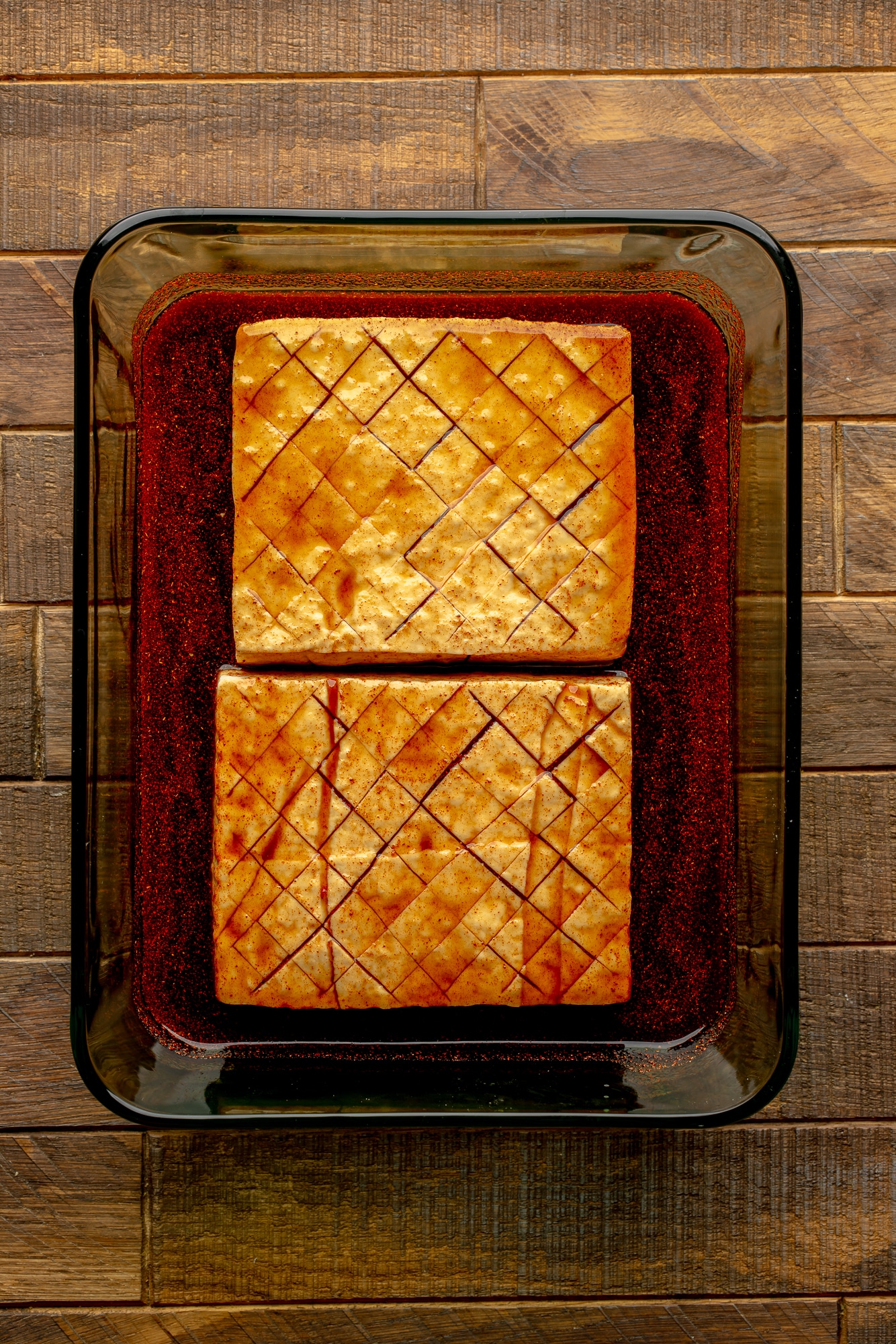 overhead view of 2 marinated and scored tofu blocks in a glass container.