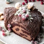 overhead view of a vegan yule log cake topped with sugared cranberries and meringue mushrooms on a white plate.