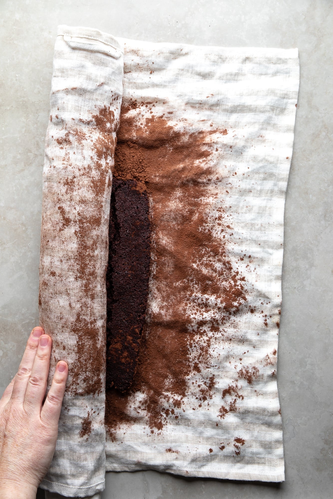 overhead view of a hand rolling a chocolate cake in a white cocoa powder-dusted towel.