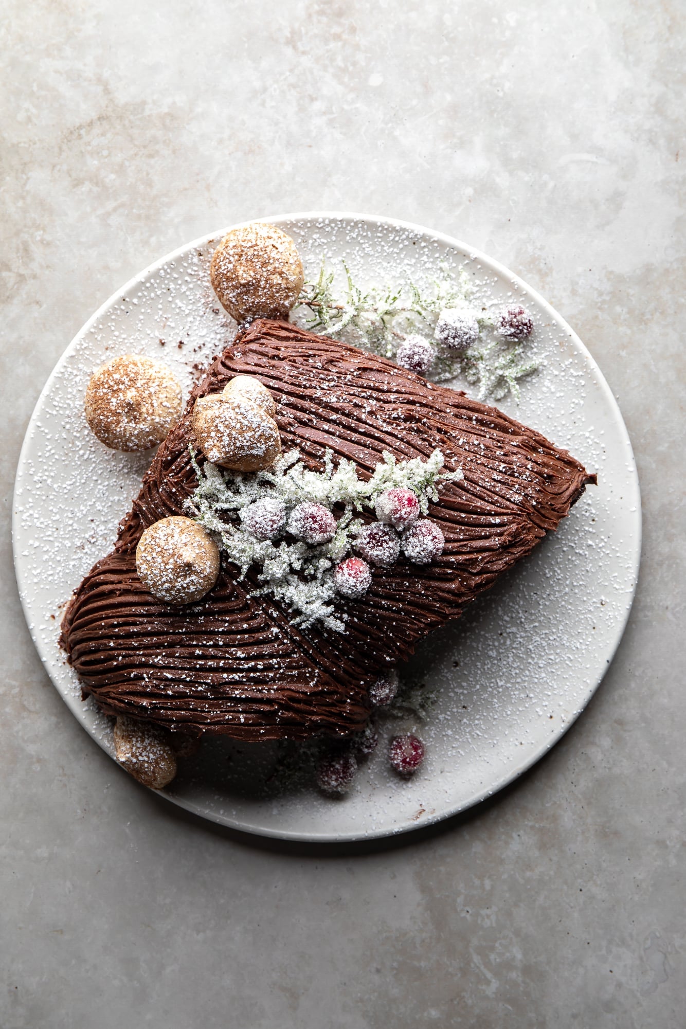 overhead view of a vegan yule log cake topped with sugared cranberries and meringue mushrooms on a white plate.