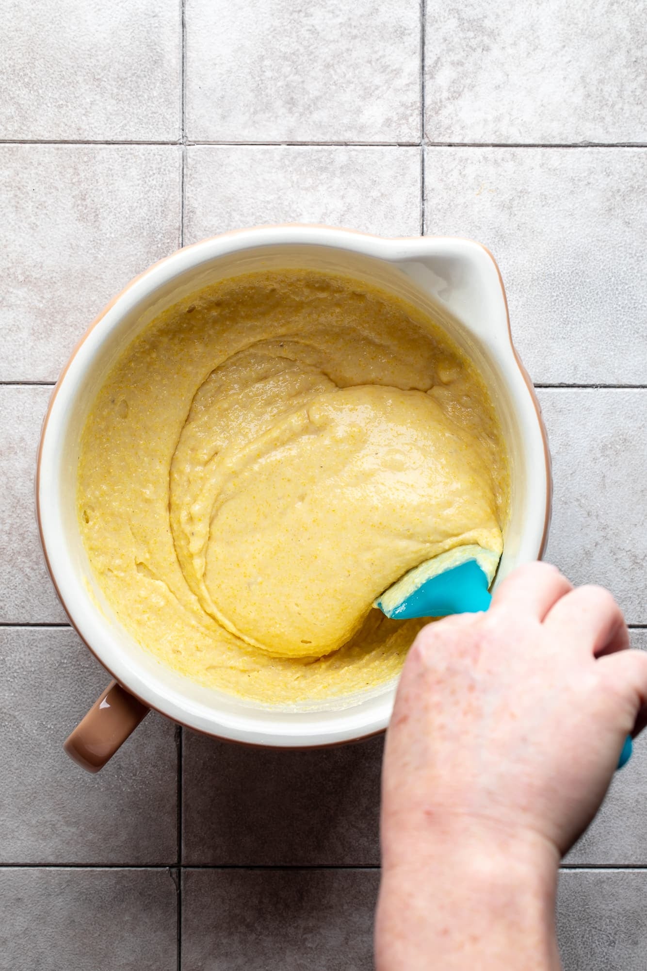 overhead view of a hand using a blue spatula to stir vegan cornbread batter in a bowl.