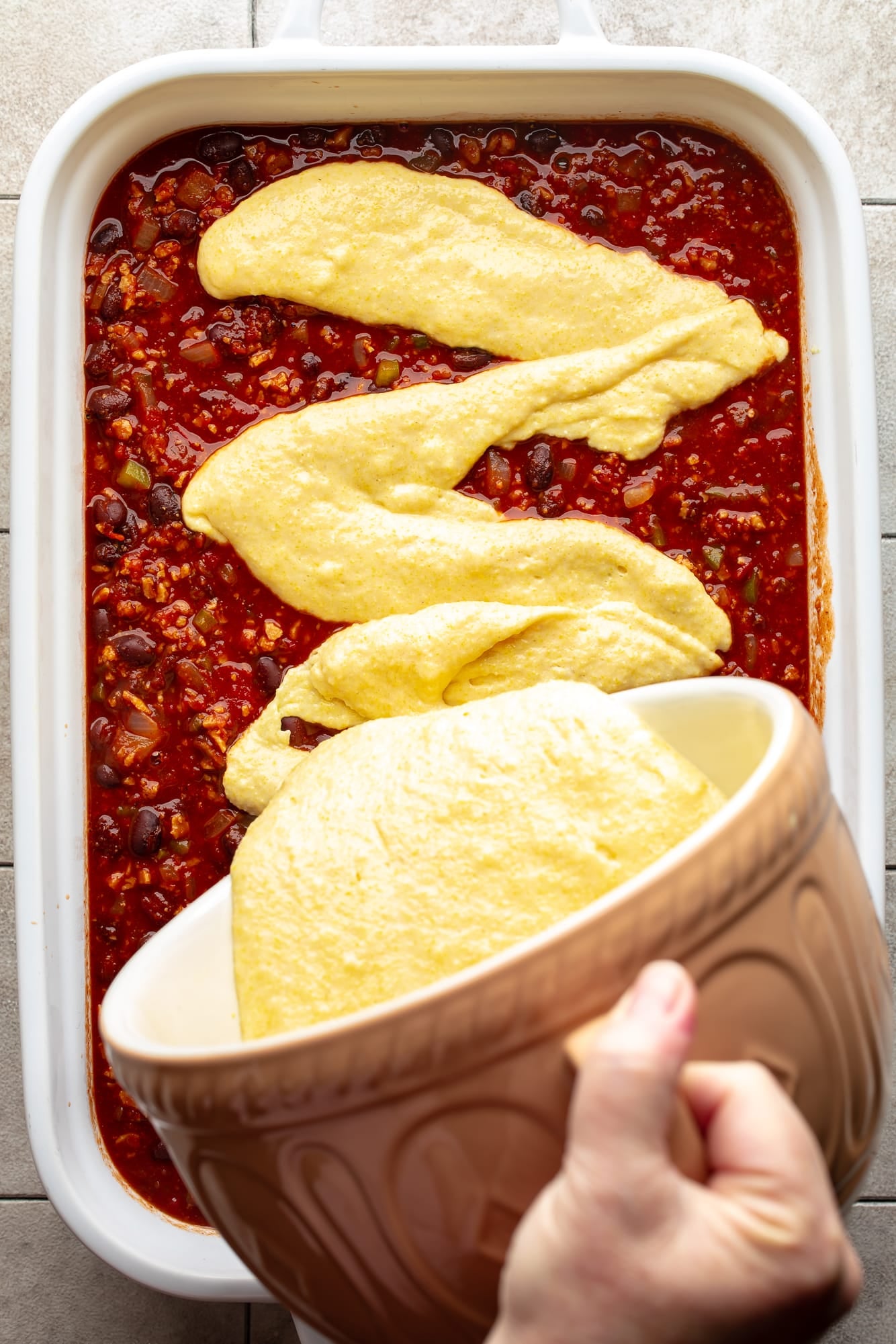 overhead view of cornbread batter being poured onto chili in a white casserole dish.