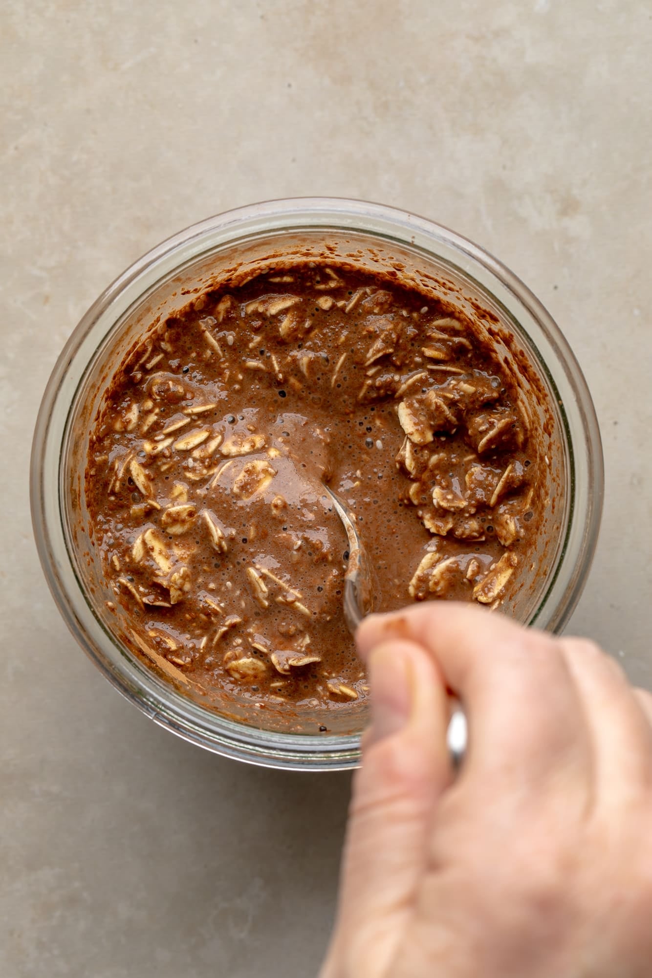overhead view of a persons hand stirring a spoon in a jar of chocolate overnight oats.