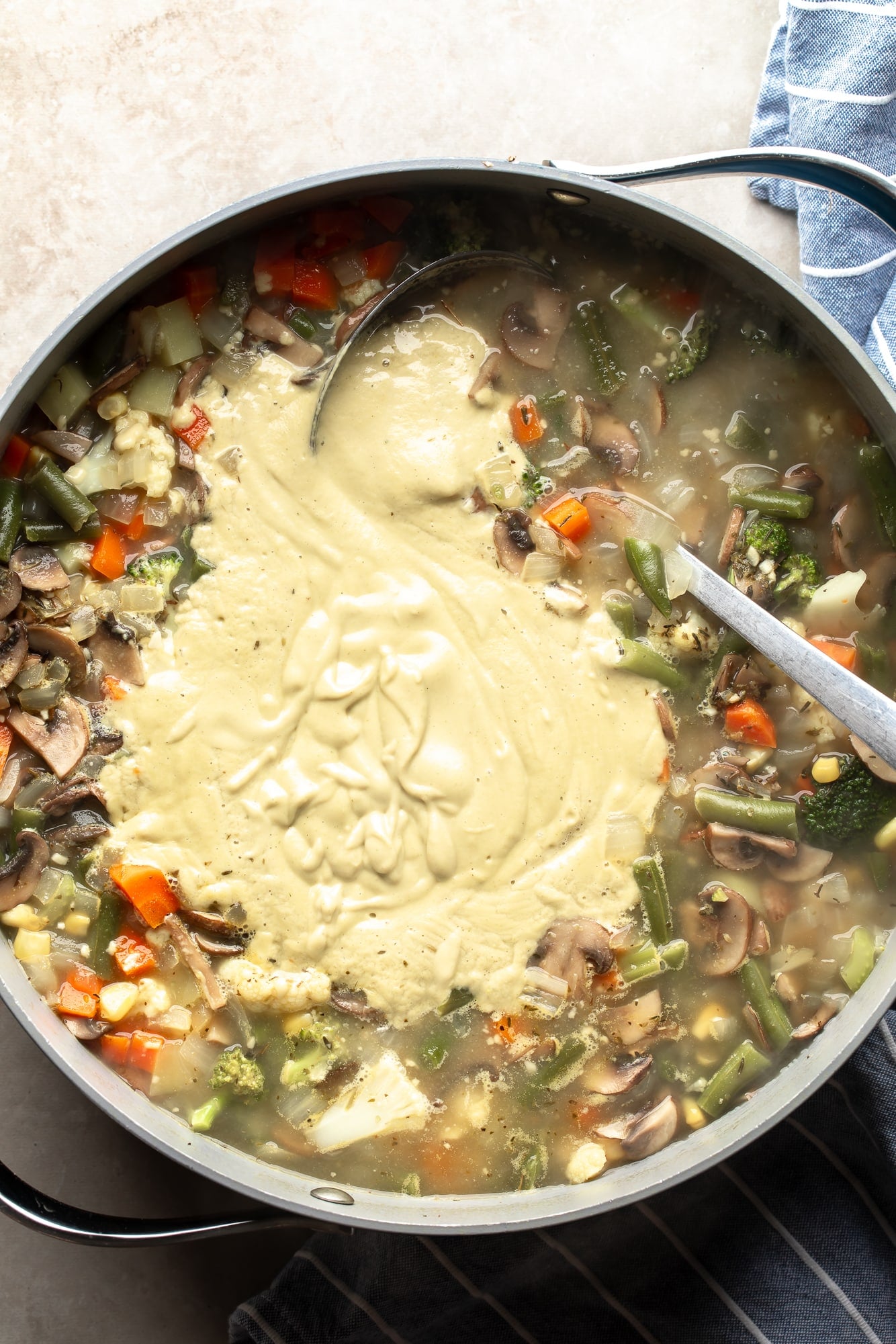 overhead view of a ladle stirring cashew cream into a large pot of Creamy Vegetable Soup.