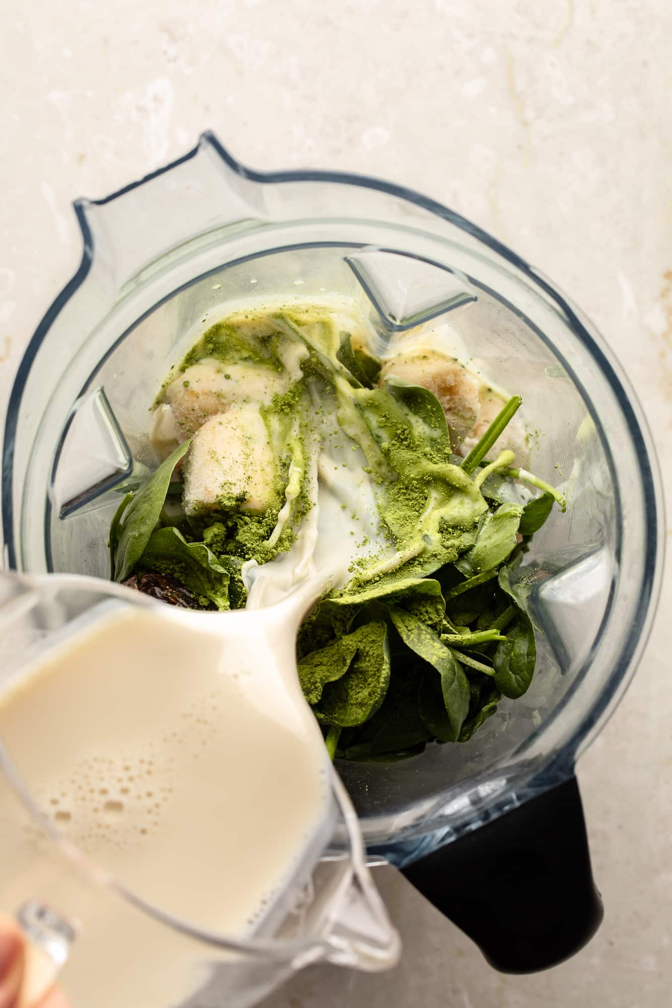overhead view of milk being poured over the ingredients for a matcha smoothie in a large blender.