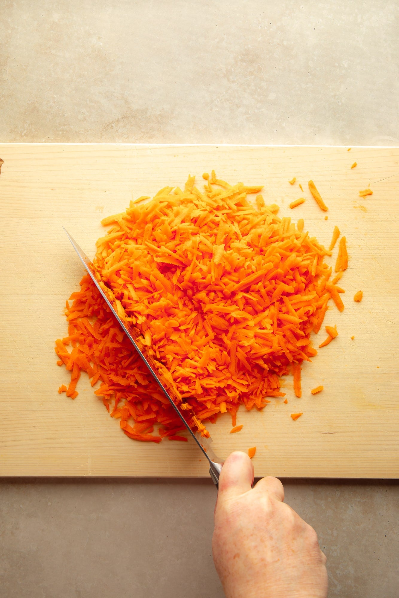 overhead view of a hand using a knife to chop grated carrots on a wood cutting board.