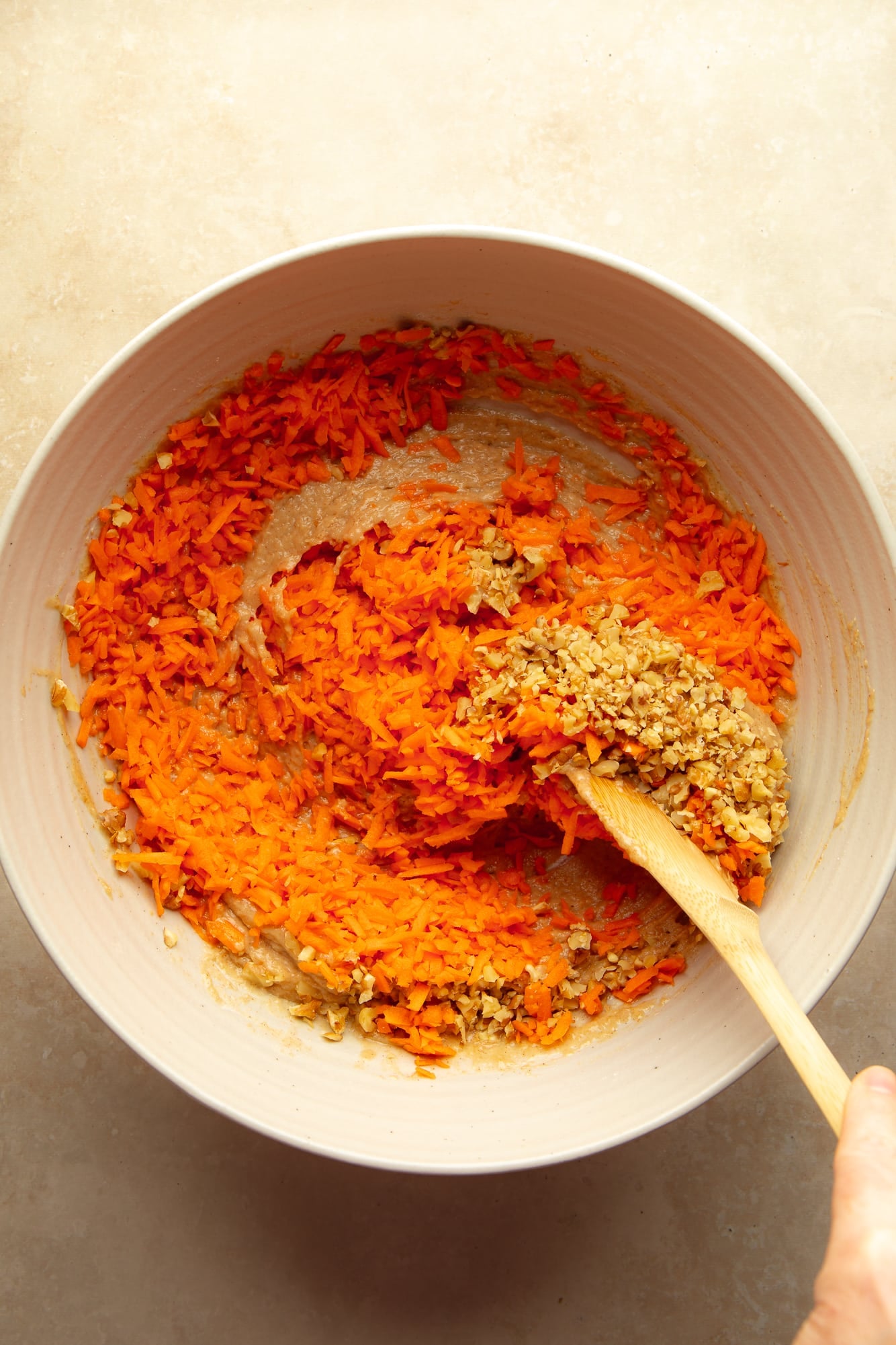overhead view of a hand using a wooden spoon to stir shredded carrots into vegan carrot cake cupcake batter in a bowl.