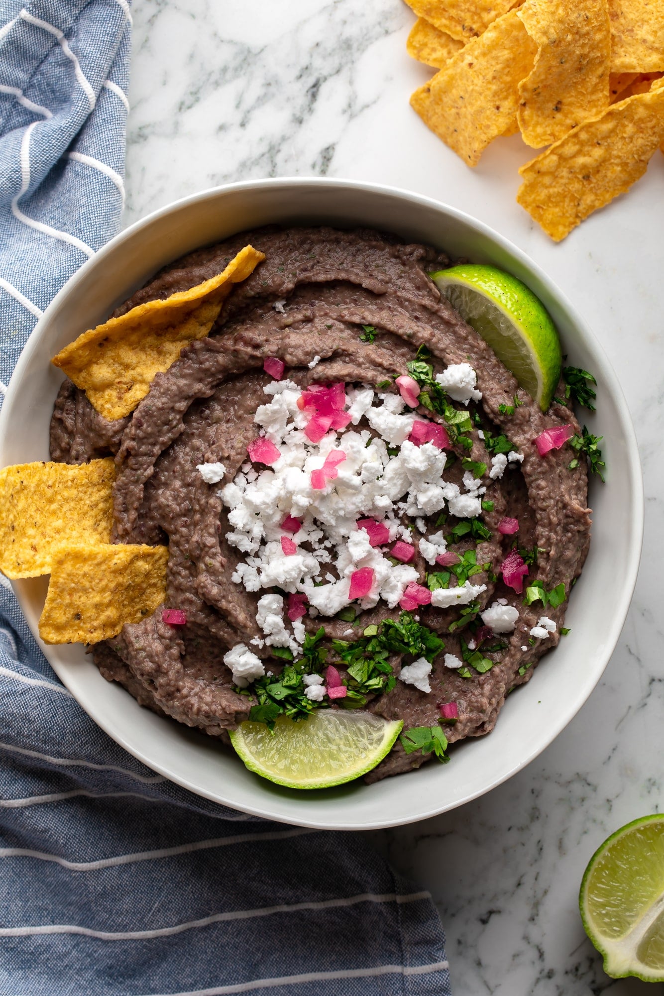 overhead view of black bean dip topped with vegan feta, cilantro, lime wedges, and tortilla chips.