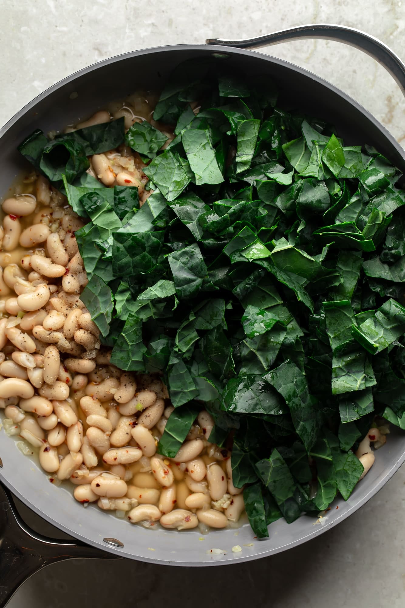 overhead view of raw kale on top of braised white beans in a skillet.
